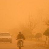 تصویری از آلودگی هوای خوزستان که در آن هوا نارنجی شده است. در تصویر یک موتور سیکلت، و دو خودرو در حال حرکتند.