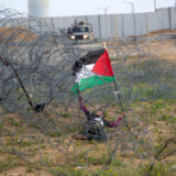 کودک فلسطینی با پرچم فلسطین در زیر سیم‌های خاردار گیرافتاده. سربازان اسرائیلی از دور نظاره‌گرند