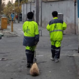 دو رفتگر نوجوان در حالی‌که یکی از آن‌ها جارویی در دست دارد از پشت سر در یک پیاده‌رو دیده می‌شوند.