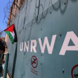 عکس یک دیوار آهنی که روی آن نوشته UNRWA. بالای دیوار سیمهای خاردار است. یک جوان فلسطینی با پرچم فلسطین به دیوار تکیه داده.