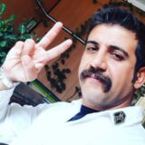 ساسان احمدی، با سبیل پهن و لباس سفید پرستاری، در حالی که دو انگشتش را به نشانه پیروزی بالا آورده و به دوربین نگاه میکند