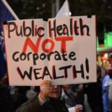 تجمعی اعتراضی در استرالیا یک نفر یک تابلو بالا گرفته که روی آن به انگلیسی نوشته: Public Health NOT Corporate Wealth (سلامت عمومی، نه ثروت شرکت‌ها)